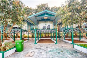 Un espacio al aire libre cubierto con una estructura tipo pérgola, rodeado de árboles y un cubo de basura verde.