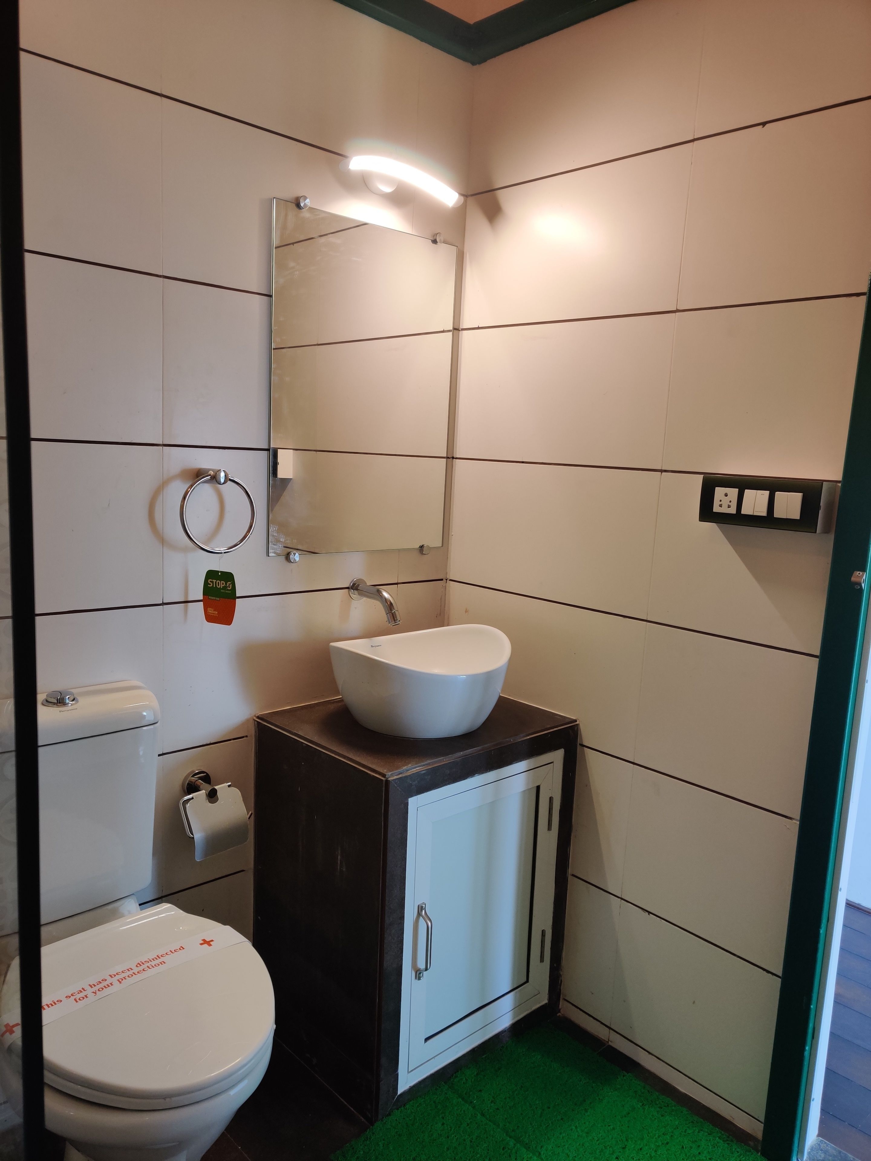 A small, clean bathroom with a white toilet, a white sink, and a mirror above it. There is a green mat on the floor, and a sign on the toilet lid.