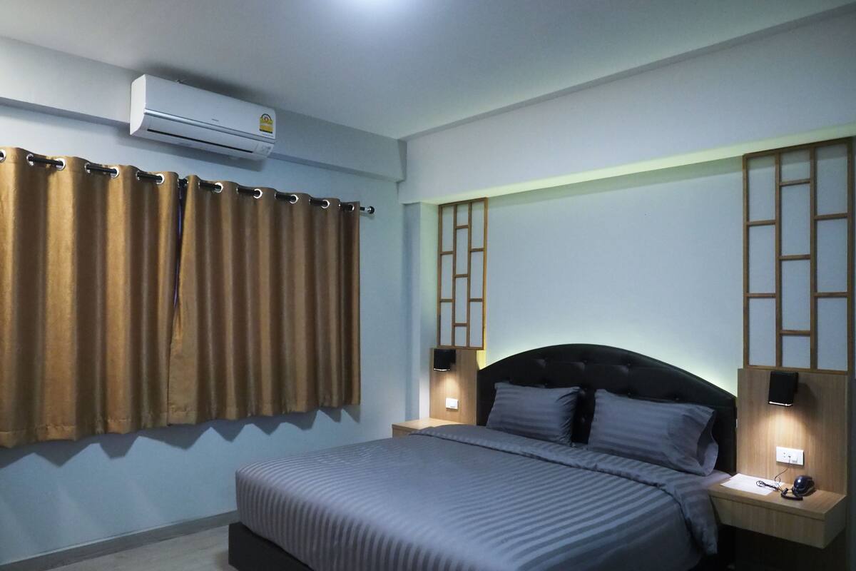 A bedroom with a bed, an air conditioner, curtains, a nightstand, and a wall-mounted shelf.