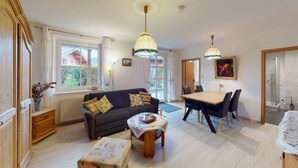 A cozy living room with a sofa, a small table, and a dining area with chairs. There are large windows allowing natural light and a view of the outside. - Ferienwohnung für 1-2 Pers., ca. 49 m²,  1 Schlafzimmer, Wohnraum, Terrasse (Bischofswiesen)
