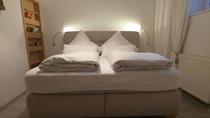 A bed with two pillows and a folded blanket, a wooden shelf with books, and a radiator. - Ferienwohnung für 1-2 Pers., ca. 49 m²,  1 Schlafzimmer, Wohnraum, Terrasse (Bischofswiesen)