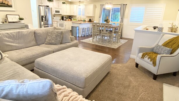 A modern living room with a grey sofa, a matching ottoman, and a white armchair. A kitchen with white cabinets and stainless steel appliances is visible in the background. - Comfortable/Cozy Home (American Fork)