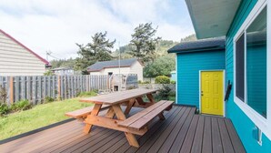 A wooden picnic table on a deck with a blue building and a yellow door in the background. - Aoy Matey · Cute family home, close to beach, fenced yard (Cloverdale)
