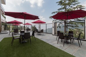 An outdoor patio with tables and chairs, red umbrellas, and a view of the mountains. - Hotel Seven Oaks (Dehradun)