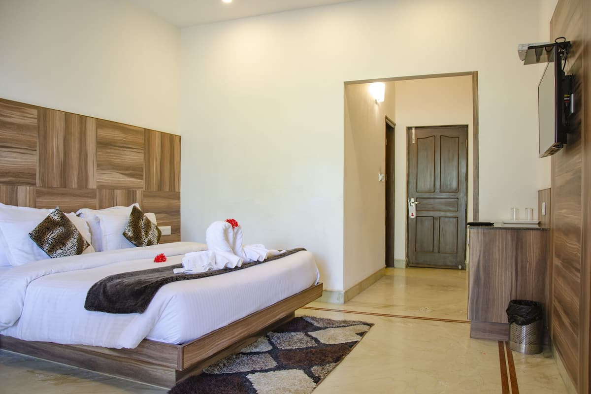 Una habitación de hotel con cabecera de madera, cama con ropa blanca, manta oscura y una pequeña alfombra. Hay un escritorio de madera con un cubo de basura y un televisor montado en la pared.