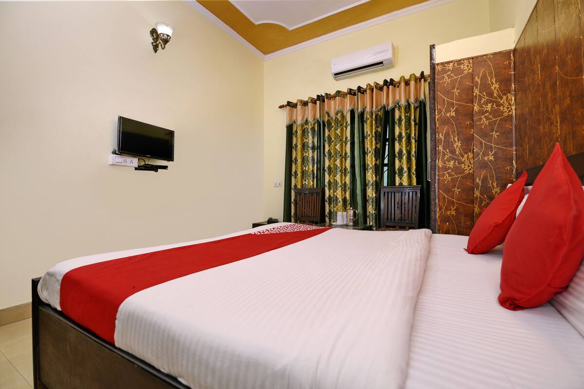 A neatly made bed with a red and white bedspread, a television mounted on the wall, and a window with curtains.