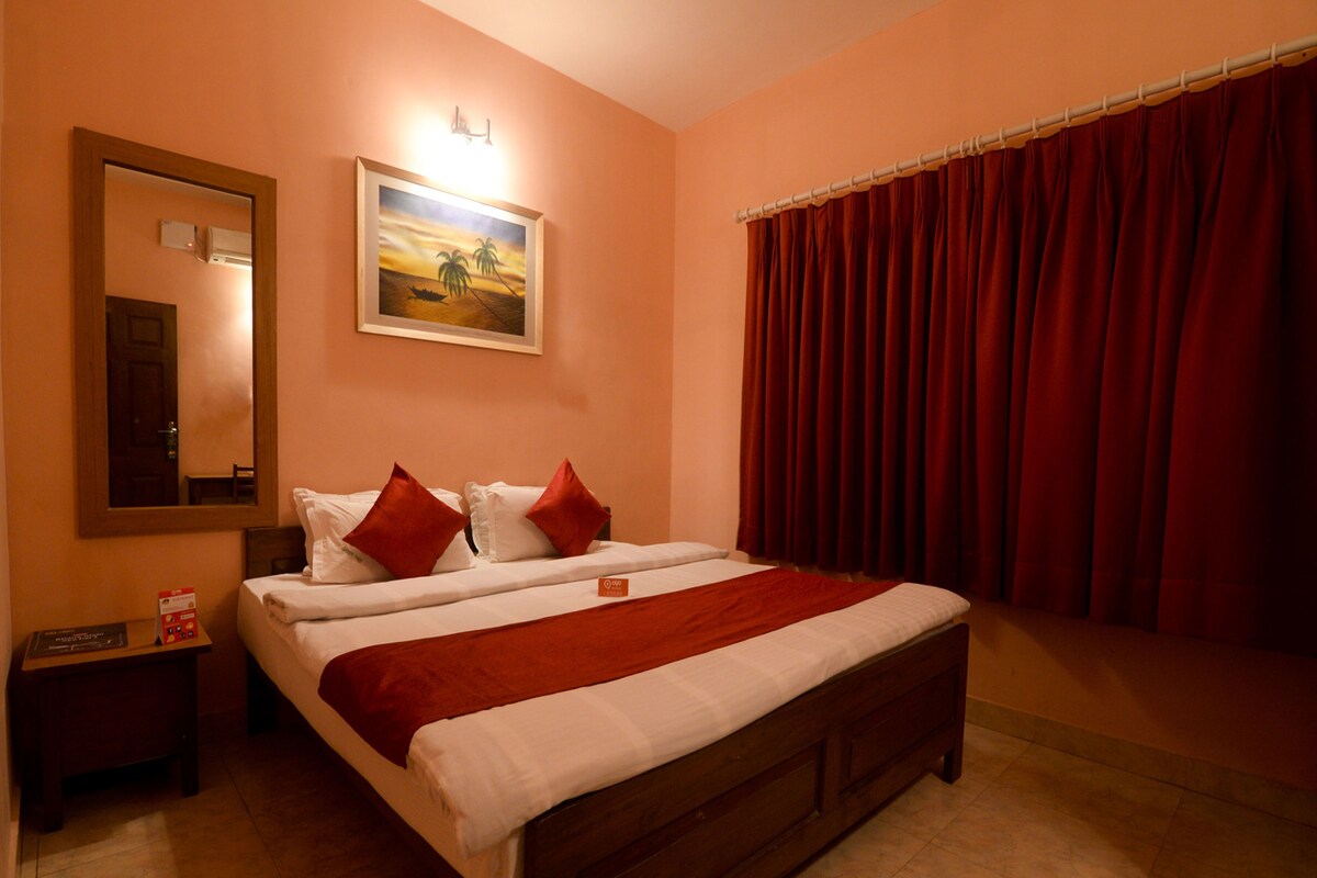 A hotel room with a wooden bed, red curtains, a painting on the wall, and a nightstand with a lamp.