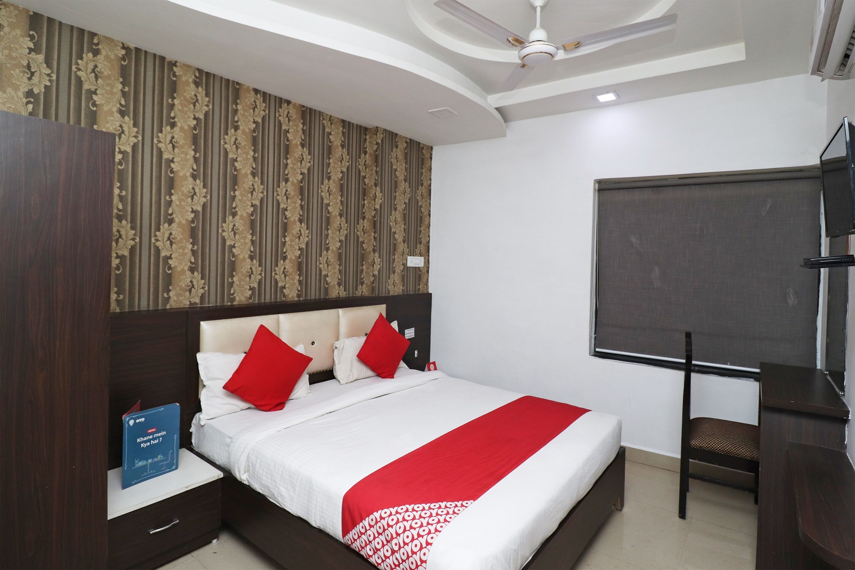 A hotel room with a bed, a desk, a chair, a TV, and a bookshelf.