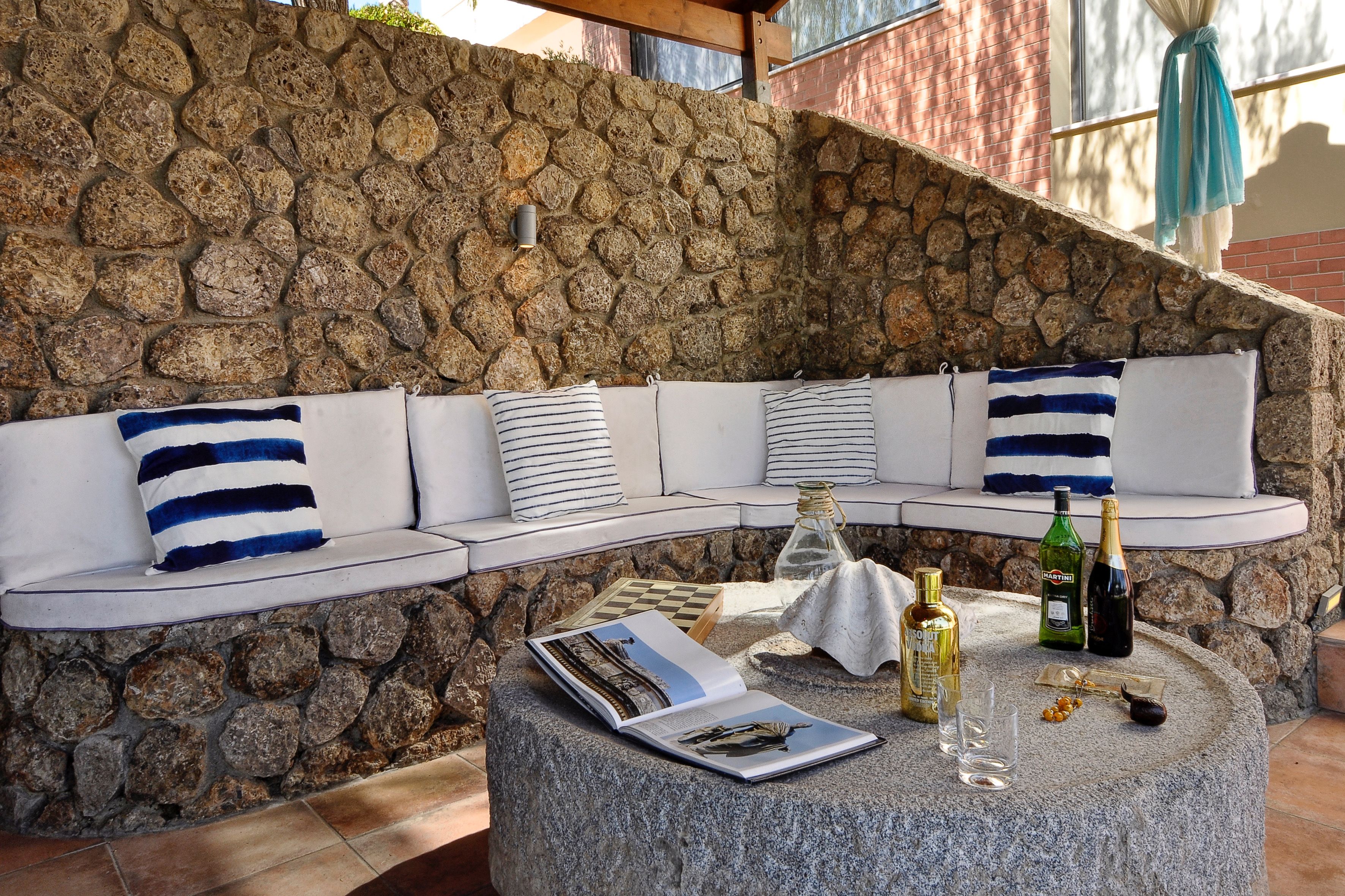 Un mur en pierre, un espace détente aux lignes courbes agrémenté de coussins à rayures, et une table ronde sur laquelle sont posés des magazines, des bouteilles et des verres.