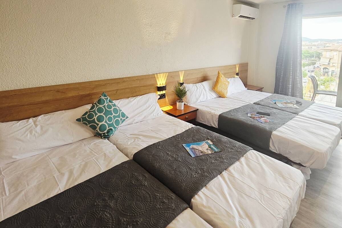Une chambre d’hôtel avec quatre lits alignés, chacun étant doté de draps blancs et de coussins décoratifs. On y trouve une tête de lit en bois, une petite table avec une lampe et une fenêtre donnant sur la ville.