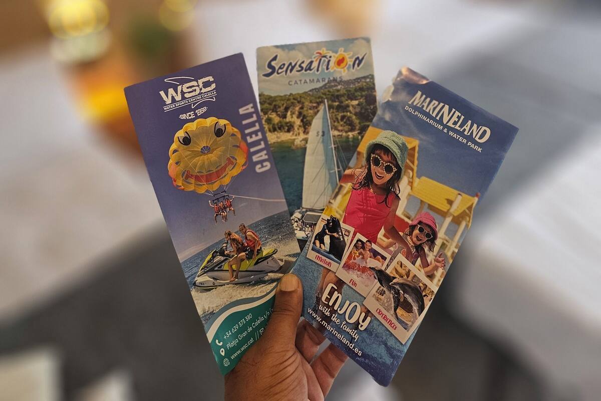 Une main tenant trois brochures de voyage illustrées de photos de sports nautiques, d’un catamaran et d’un parc aquatique avec des dauphins.