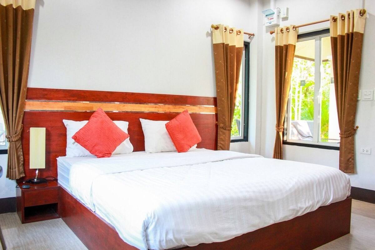 A neatly made bed with white bedding and two red pillows, a wooden headboard, a bedside table with a lamp, and curtains on the windows.