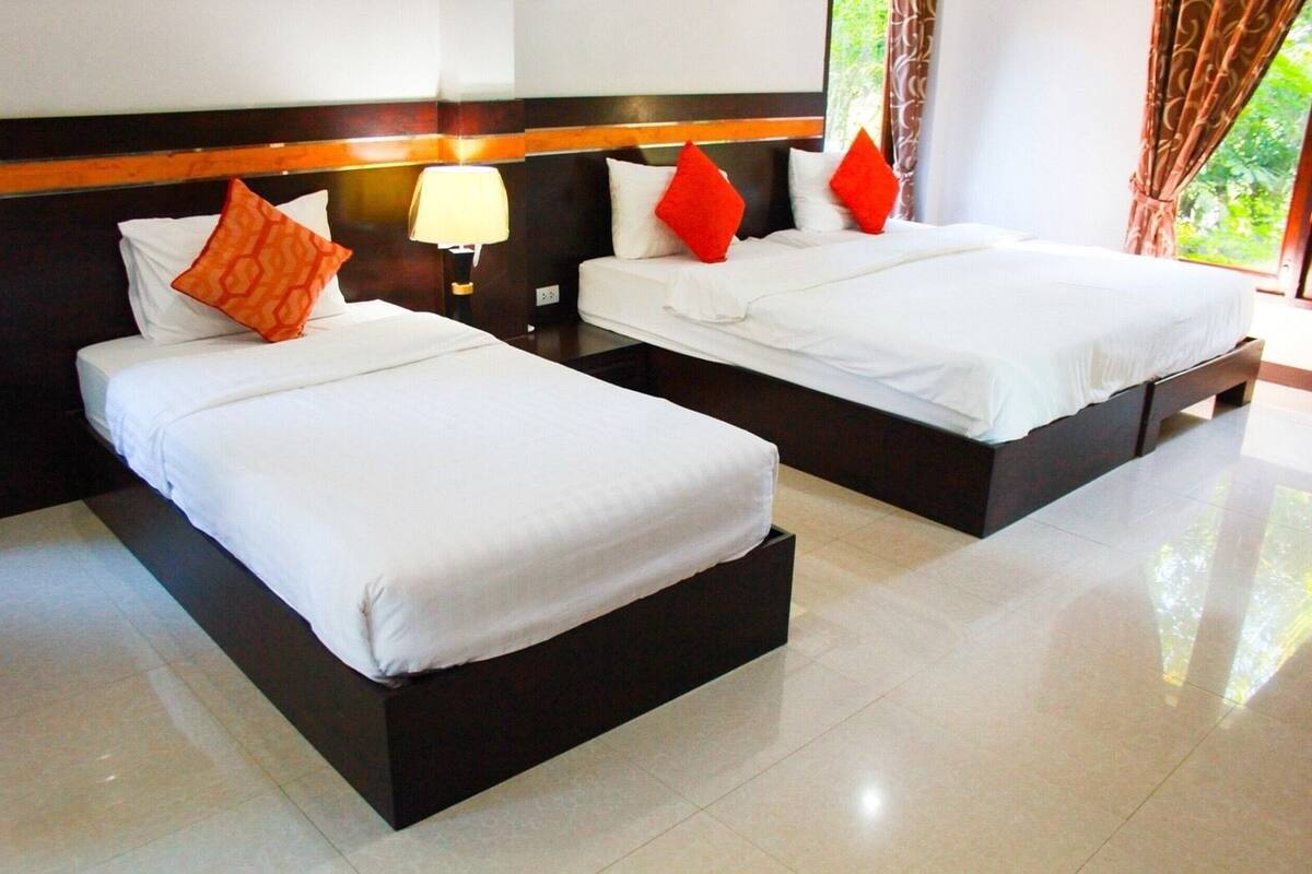 Two single beds with white linens and red pillows, a nightstand with a lamp, and a view of greenery through a window.