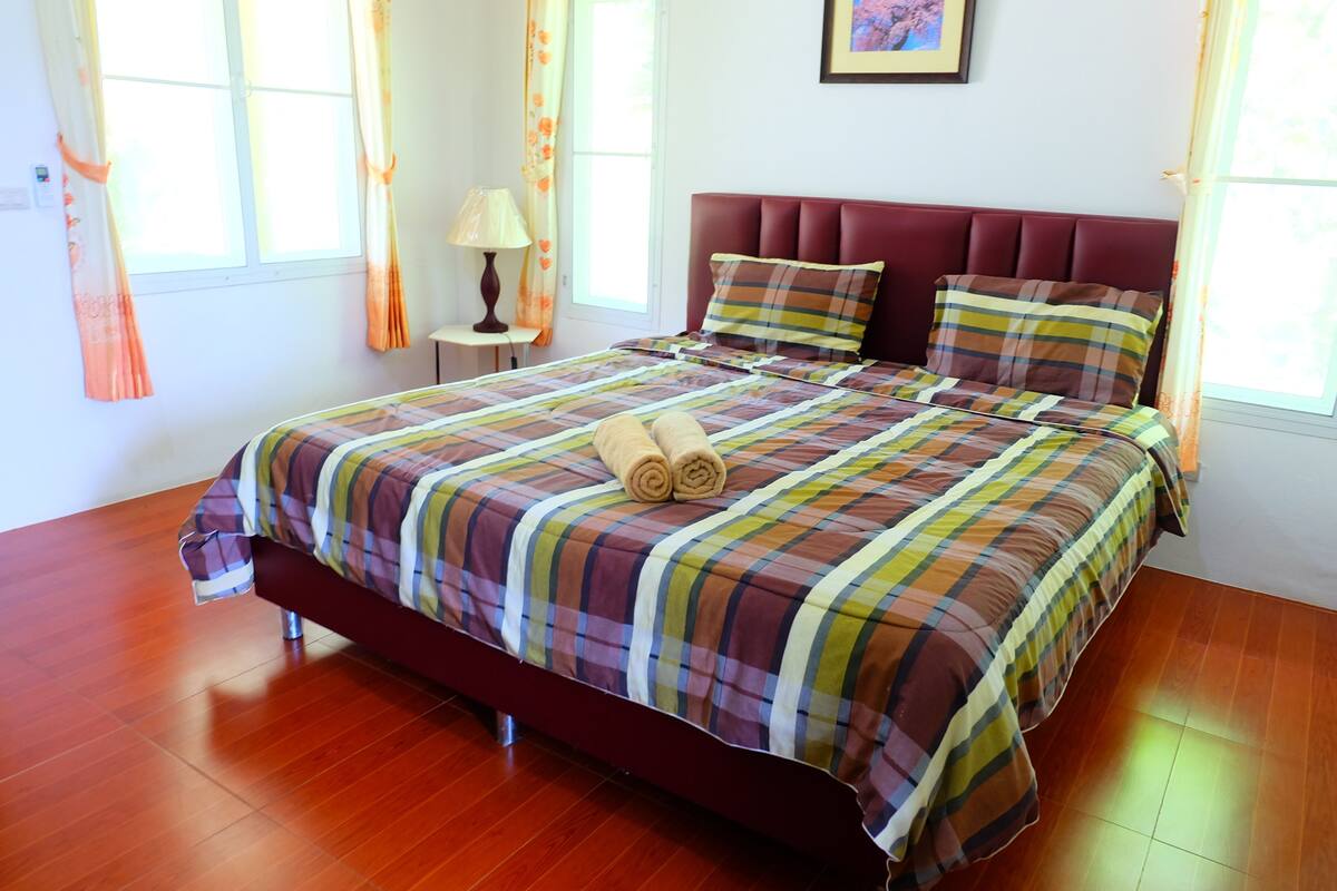A neatly made bed with a plaid bedspread and two pillows, a wooden floor, a nightstand with a lamp, and a window with curtains.