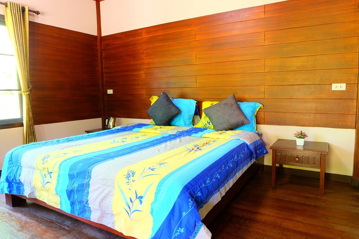 A bedroom with a wooden headboard, a bed with a blue and yellow floral bedspread, two pillows, and a small wooden nightstand with a plant.