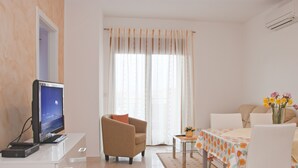 Ein modernes Wohnzimmer mit einem Flachbildfernseher, einem beigefarbenen Sessel, einem Esstisch mit einer blumigen Tischdecke und einer Couch.