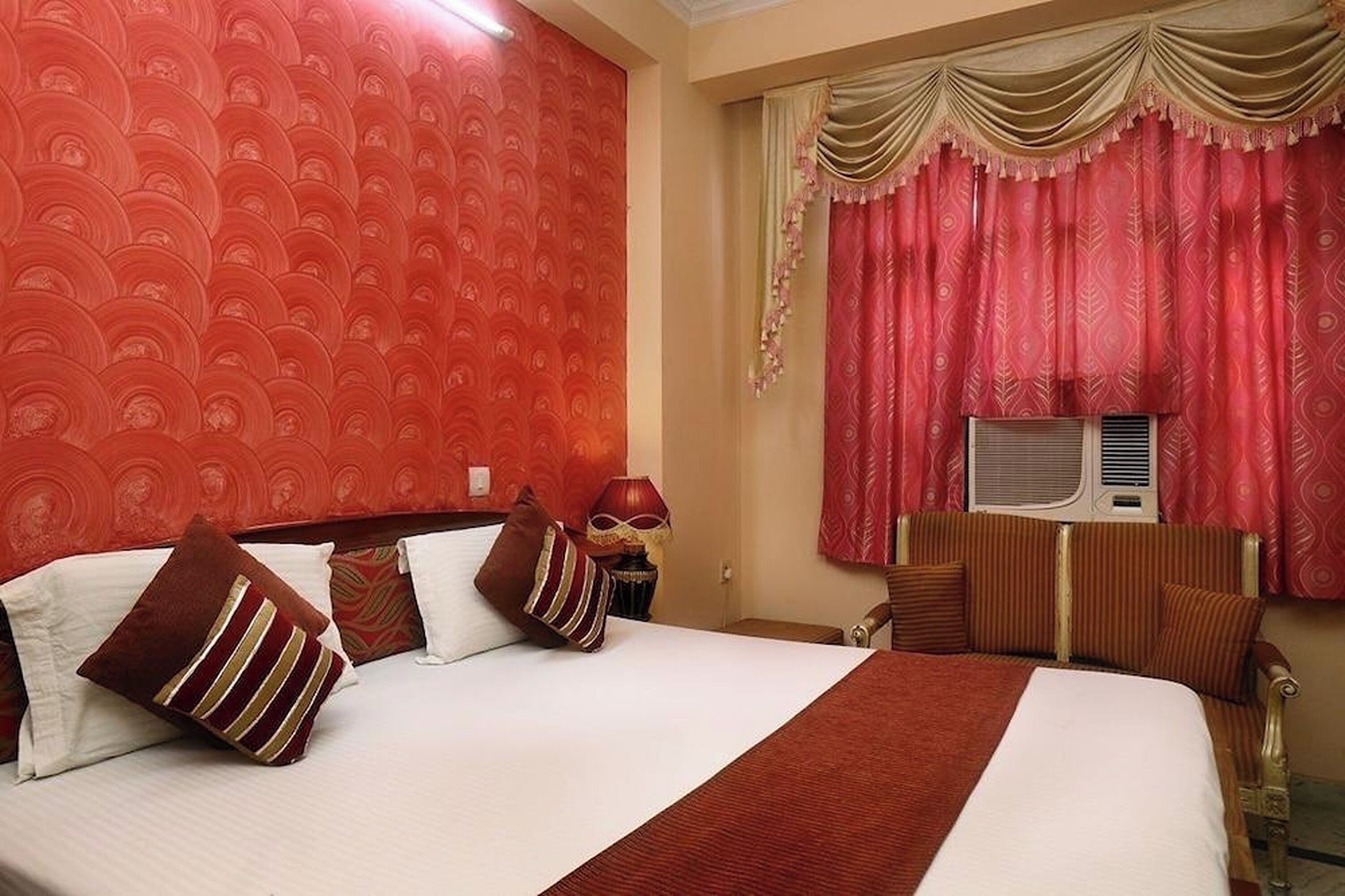 A hotel room with a large bed, red patterned wallpaper, a window with red curtains, and an air conditioning unit.