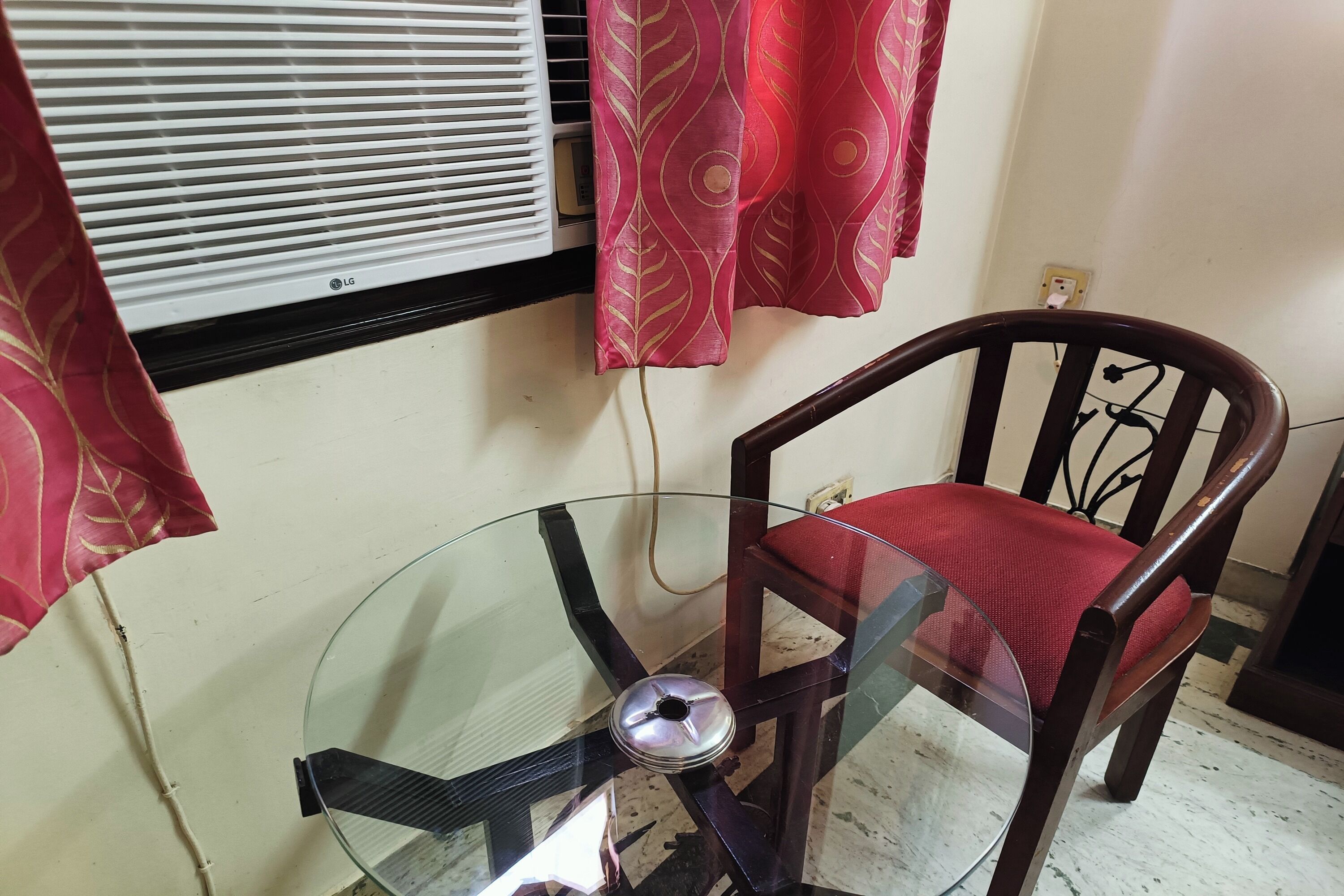 A glass-top table with a chair, a window with blinds, and a power outlet on the wall.