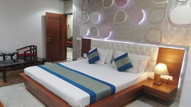 A neatly made bed with a headboard, a bedside table with a lamp, and a wooden chair. - Hotel Indraprastha (New Delhi)