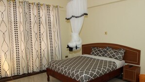 A bedroom with a bed, curtains, a bedside table, and a nightstand. - Gees Inn (Nakuru)