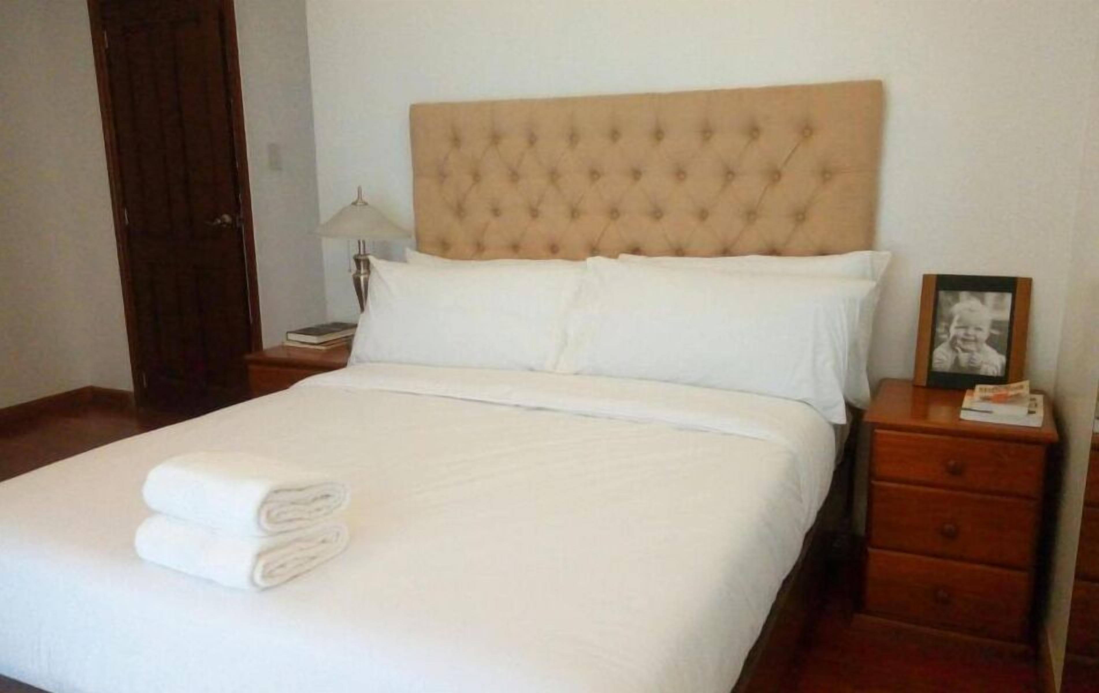 Una cama bien hecha con sábanas blancas y una toalla doblada, un cabecero de madera, una mesita de noche con una lámpara y un cuadro en el tocador.