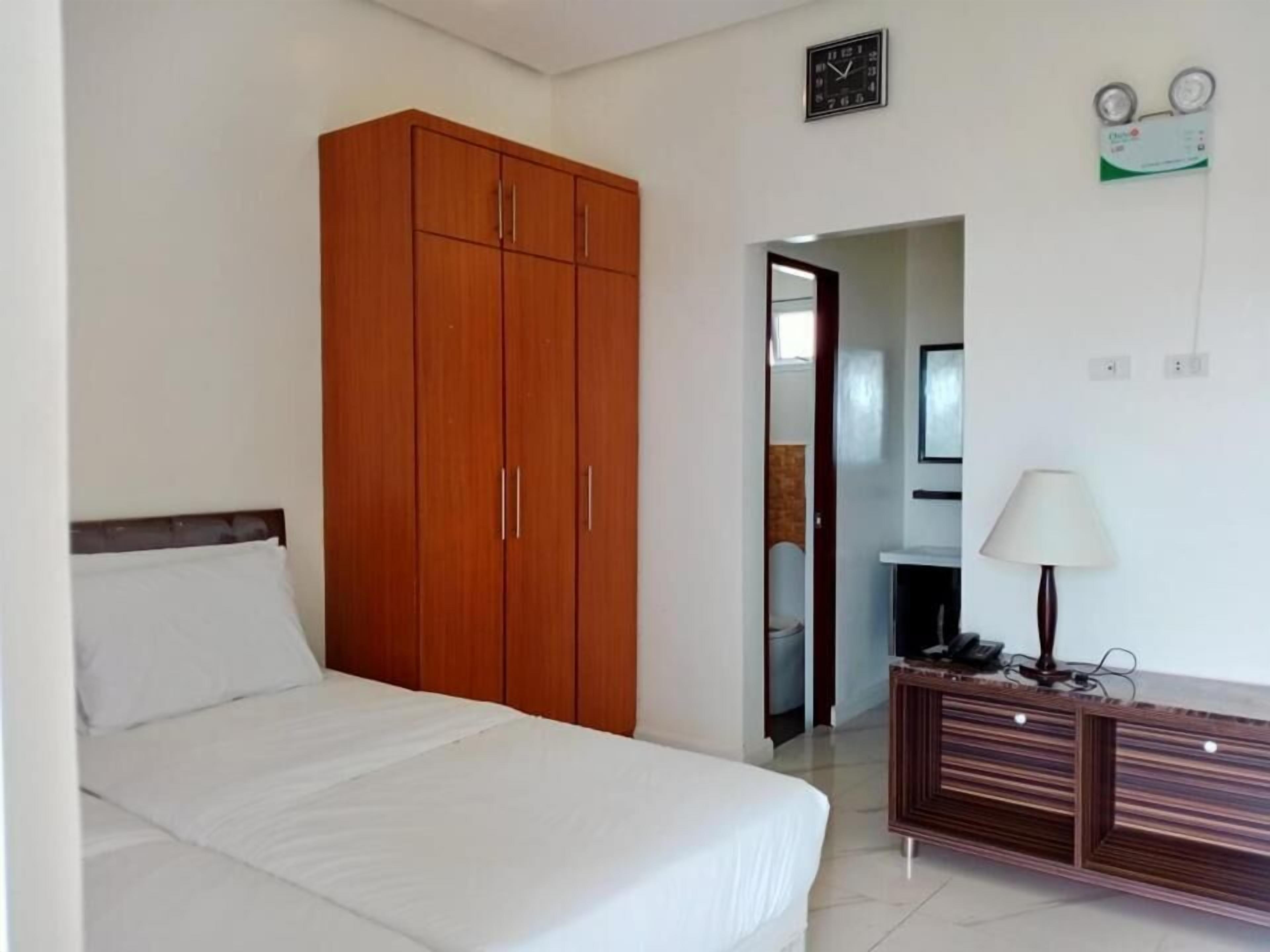 A bedroom with a large wooden wardrobe, a bed with white bedding, a bedside table with a lamp, and a clock on the wall.
