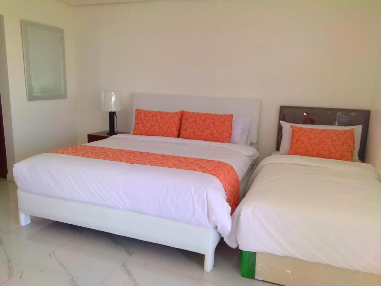 A double bed with white and orange bedding, a nightstand with a lamp, and a small green bottle on the floor.