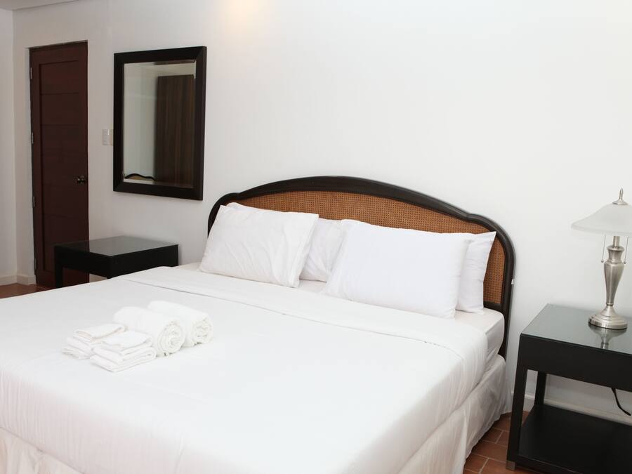 A neatly made bed with white linens and towels, a bedside table with a lamp, a mirror on the wall, and a door to the left.