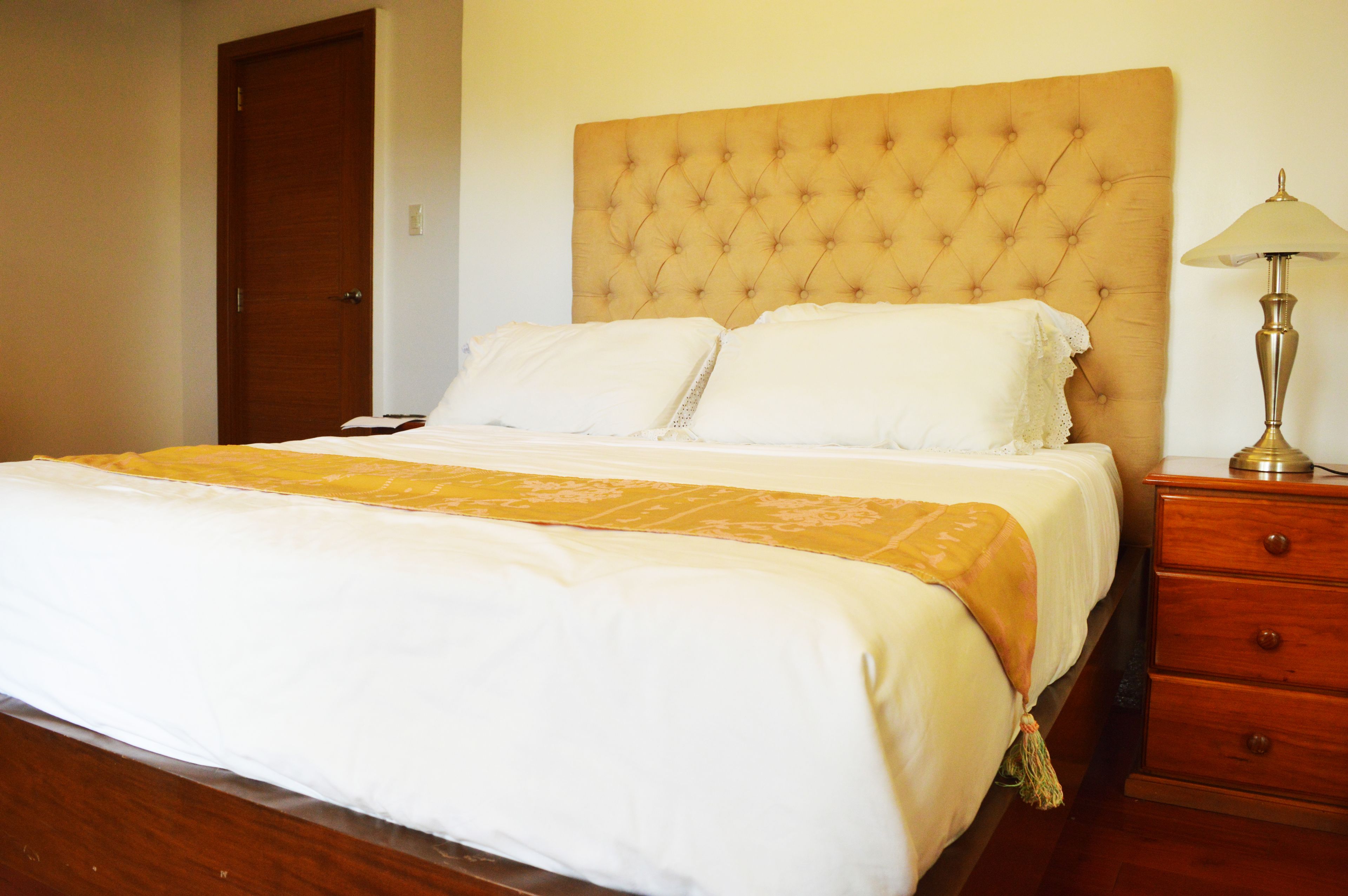 Una cama bien hecha con cabecera, lámpara de noche y mesita de madera.