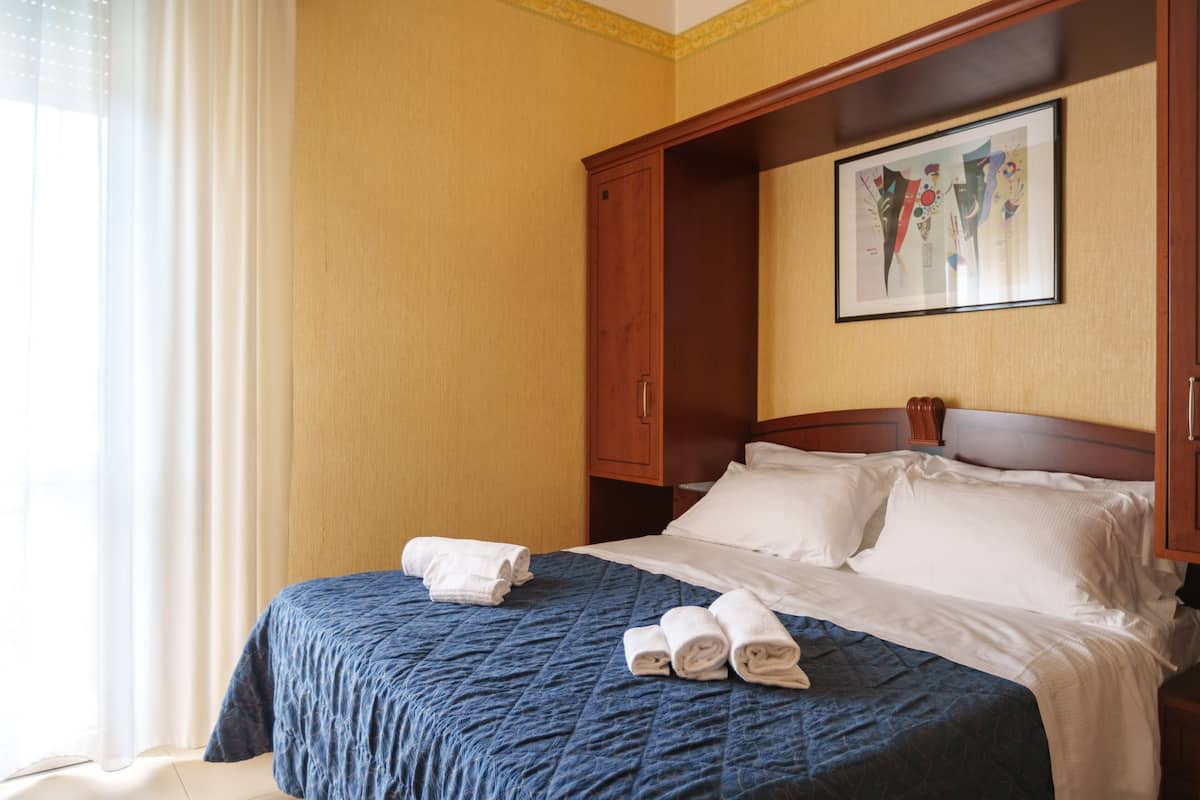 Habitación de hotel con una cama grande, un armario de madera, un cuadro en la pared y una ventana con cortinas.