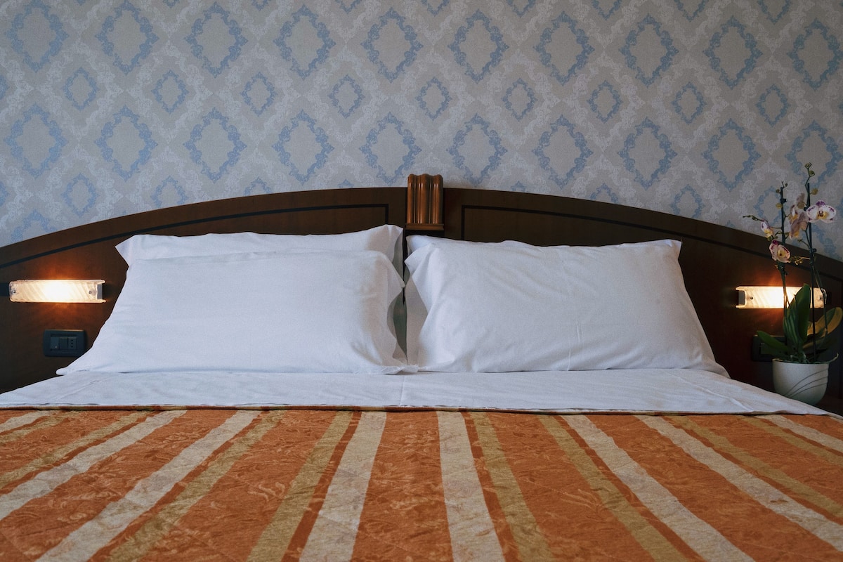 Una cama bien hecha con almohadas blancas y una colcha a rayas anaranjadas, frente a una pared con papel tapiz de diseño azul y blanco.