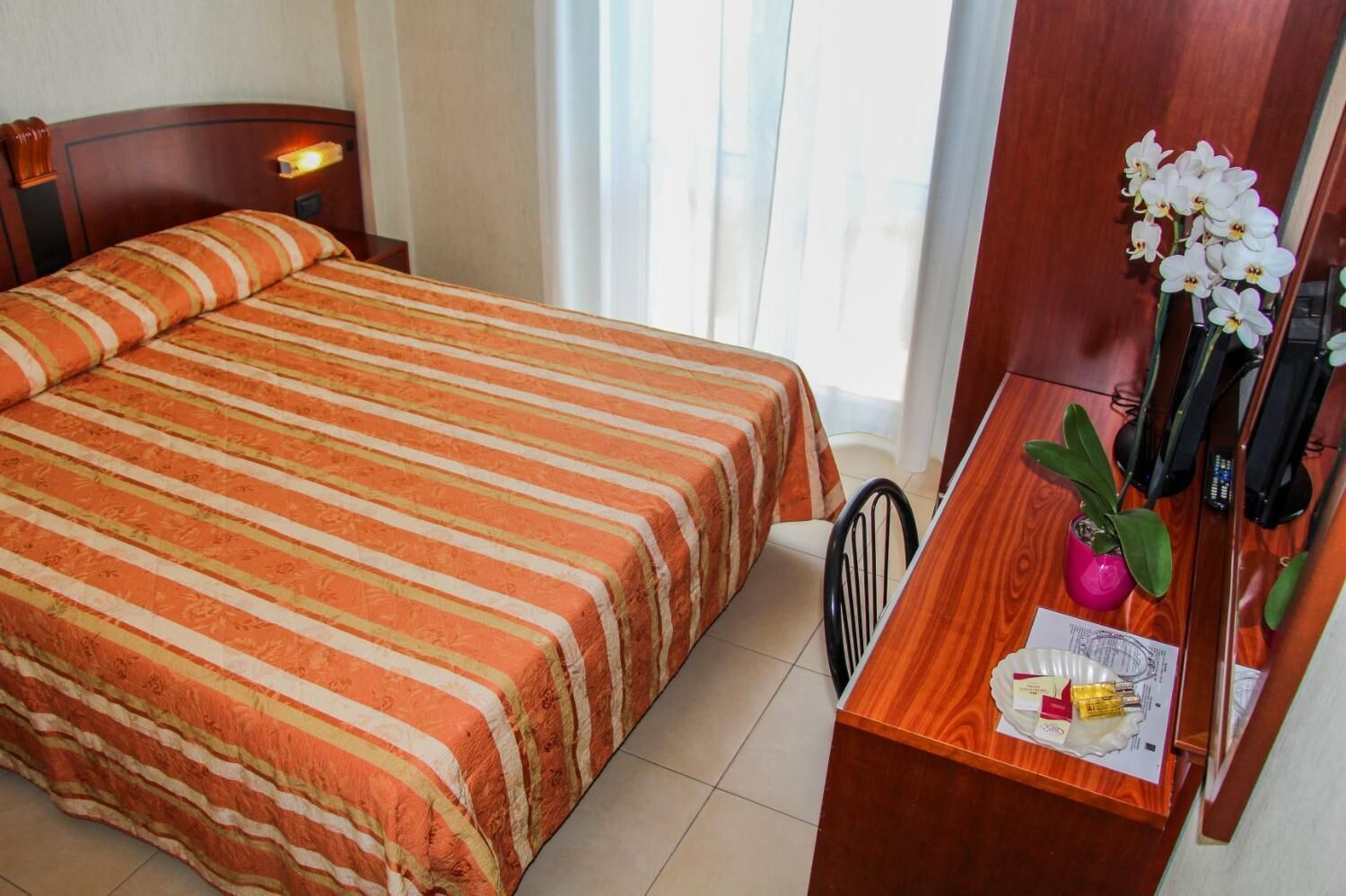 A hotel room with a bed, a desk with a vase of flowers, and a mirror.