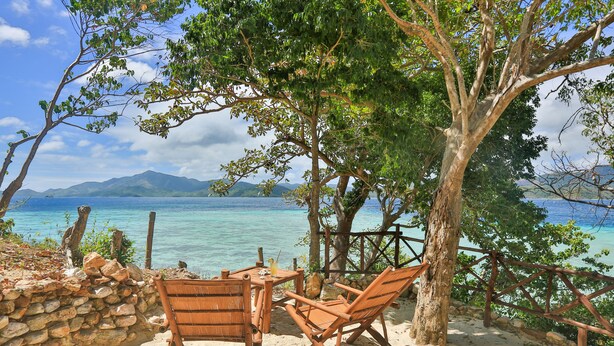 A wooden table and chairs set on a sandy beach with a view of the sea and mountains. - Bamboo Private Islands (Culion Island)