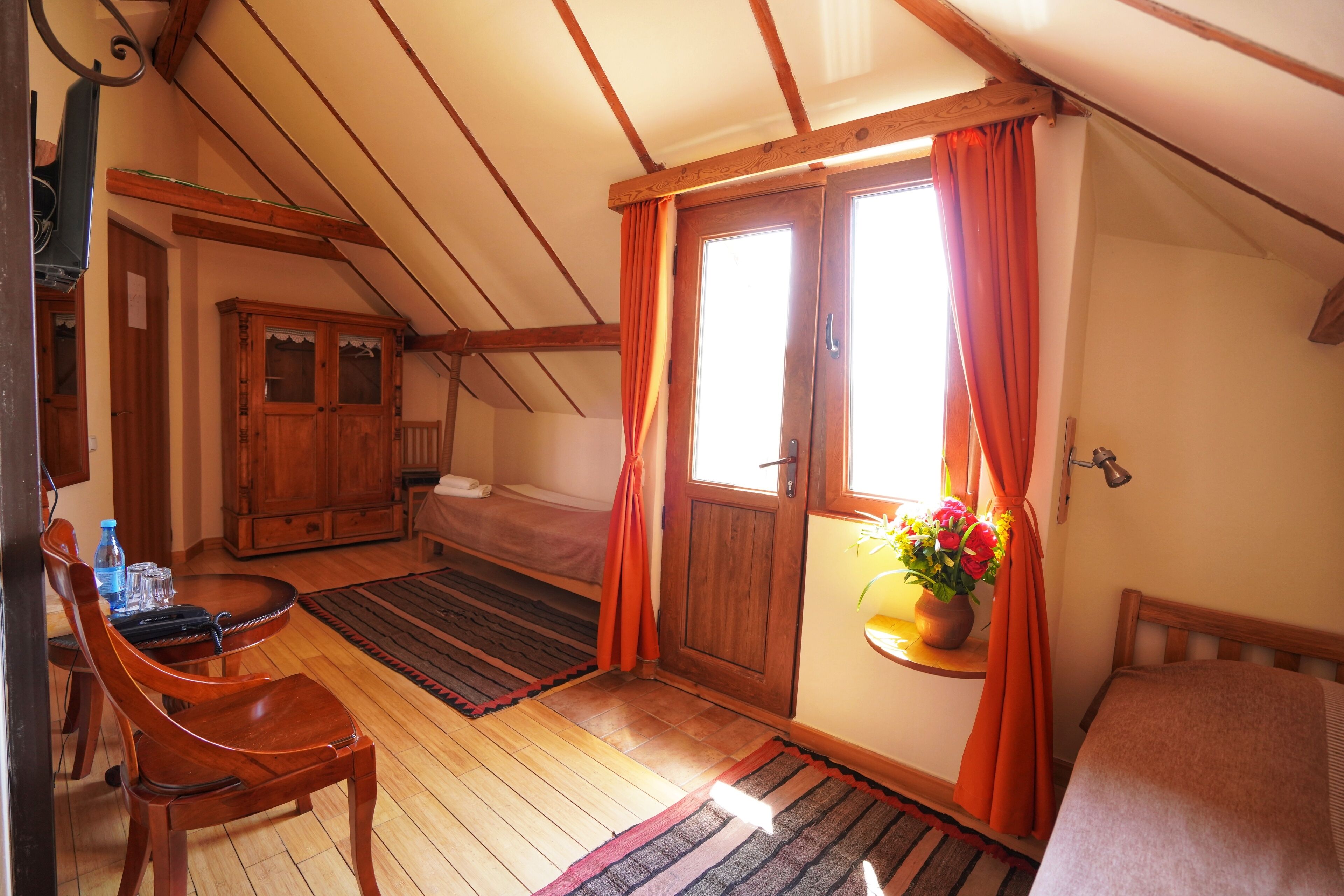 Una habitación con piso de madera, una cama, un armario de madera, una mesa con sillas y una ventana con cortinas.