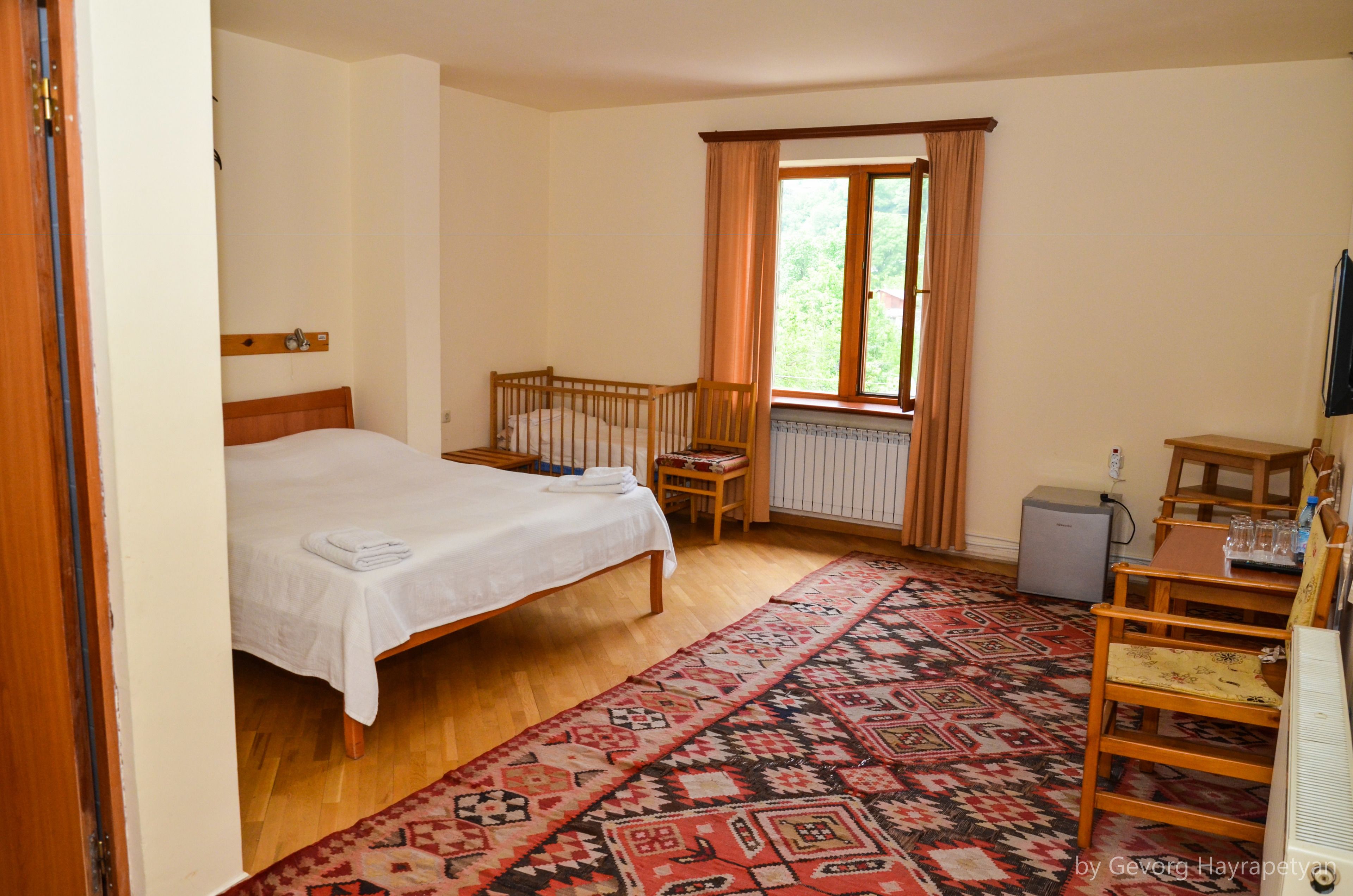 A bedroom with a bed, a wooden crib, a window with curtains, a radiator, a desk with a chair, and a patterned rug.