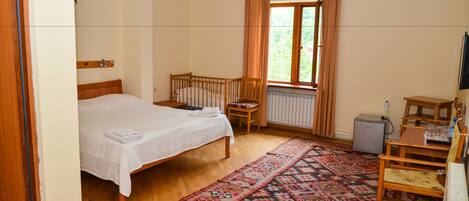 A bedroom with a bed, a wooden crib, a window with curtains, a radiator, a desk with a chair, and a patterned rug.