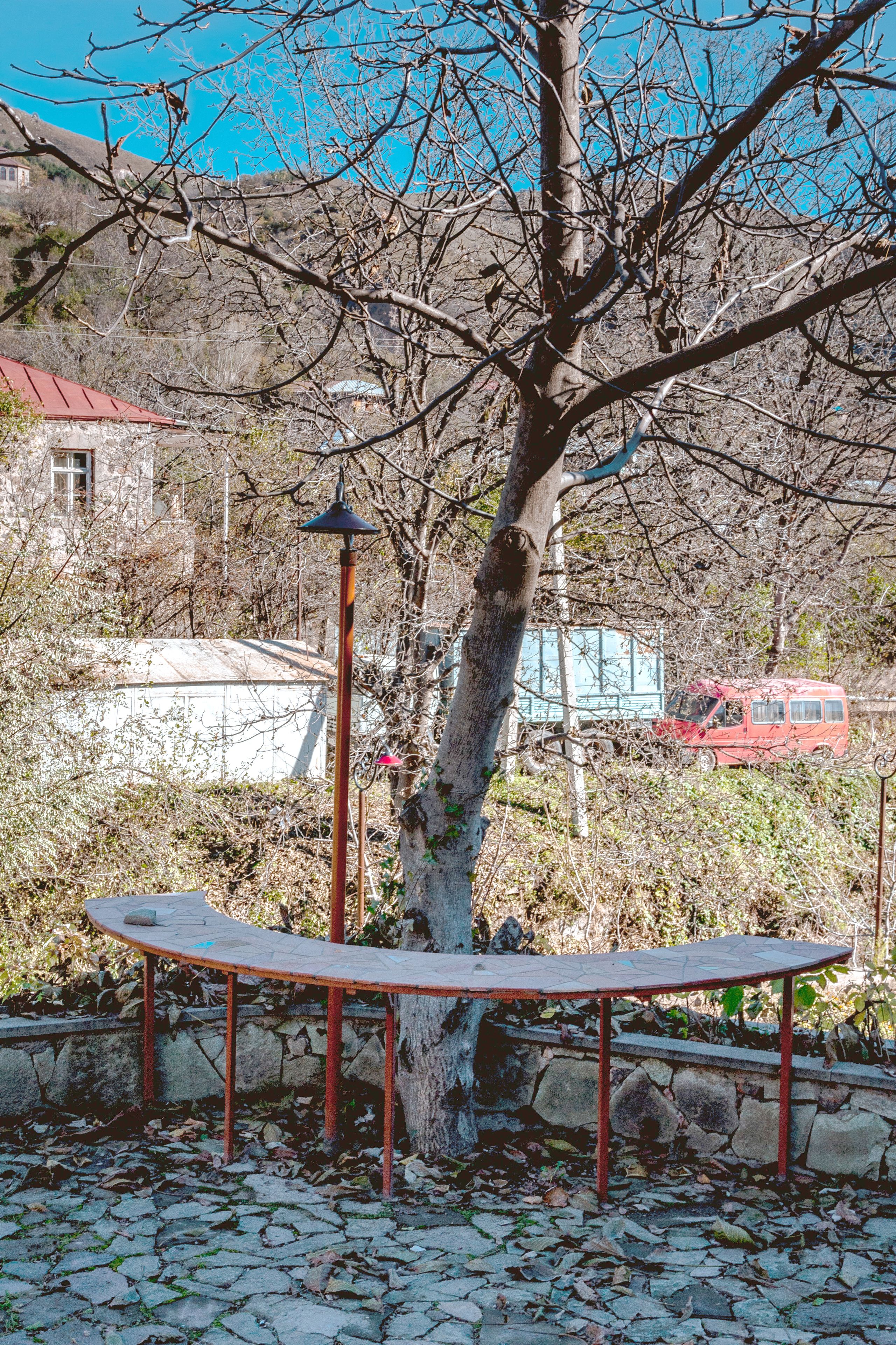 Un área empedrada con un banco, un árbol y una camioneta roja al fondo.