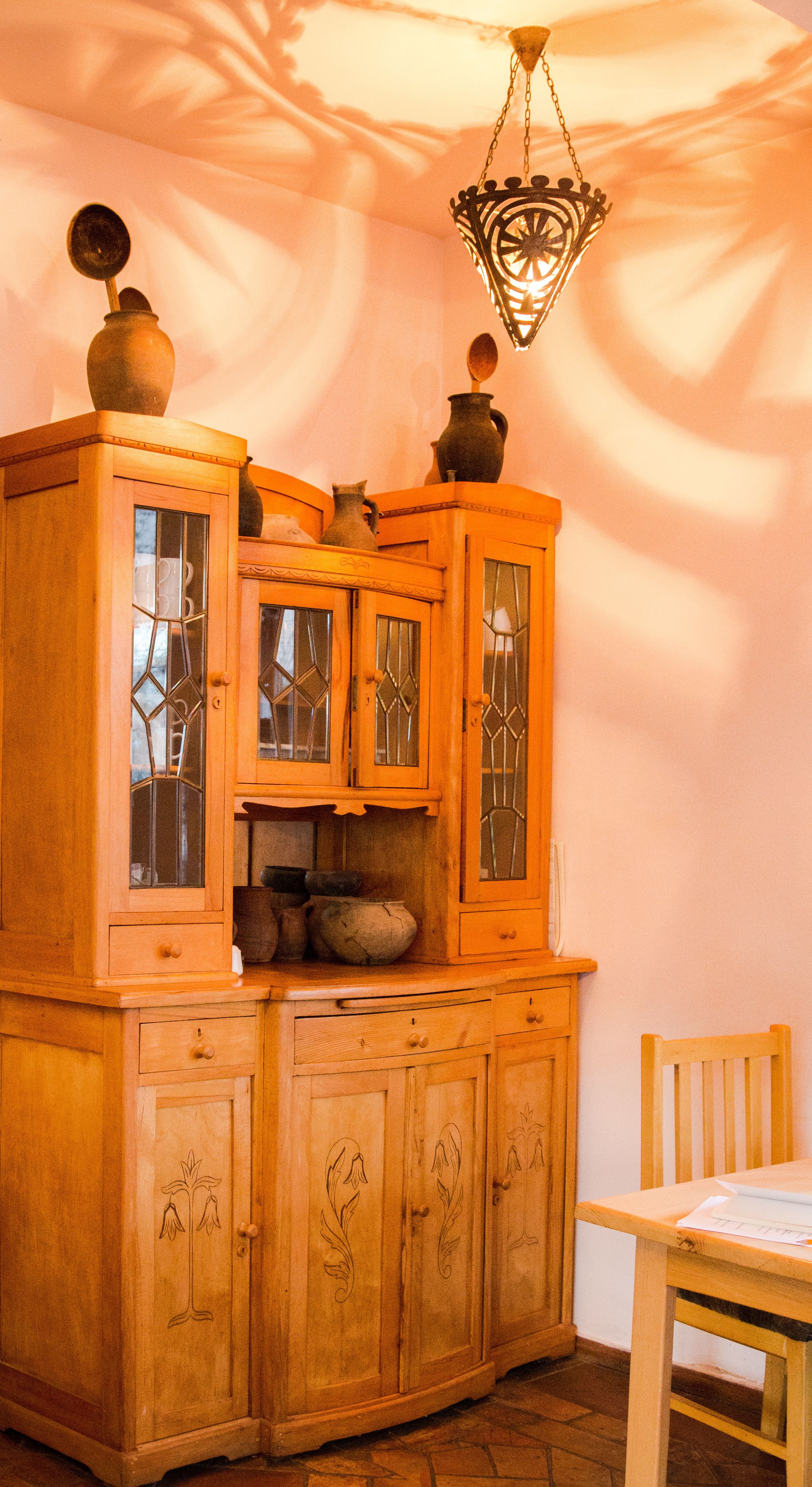 Un armario de madera con puertas de cristal y detalles decorativos, una lámpara colgante y una silla de madera.