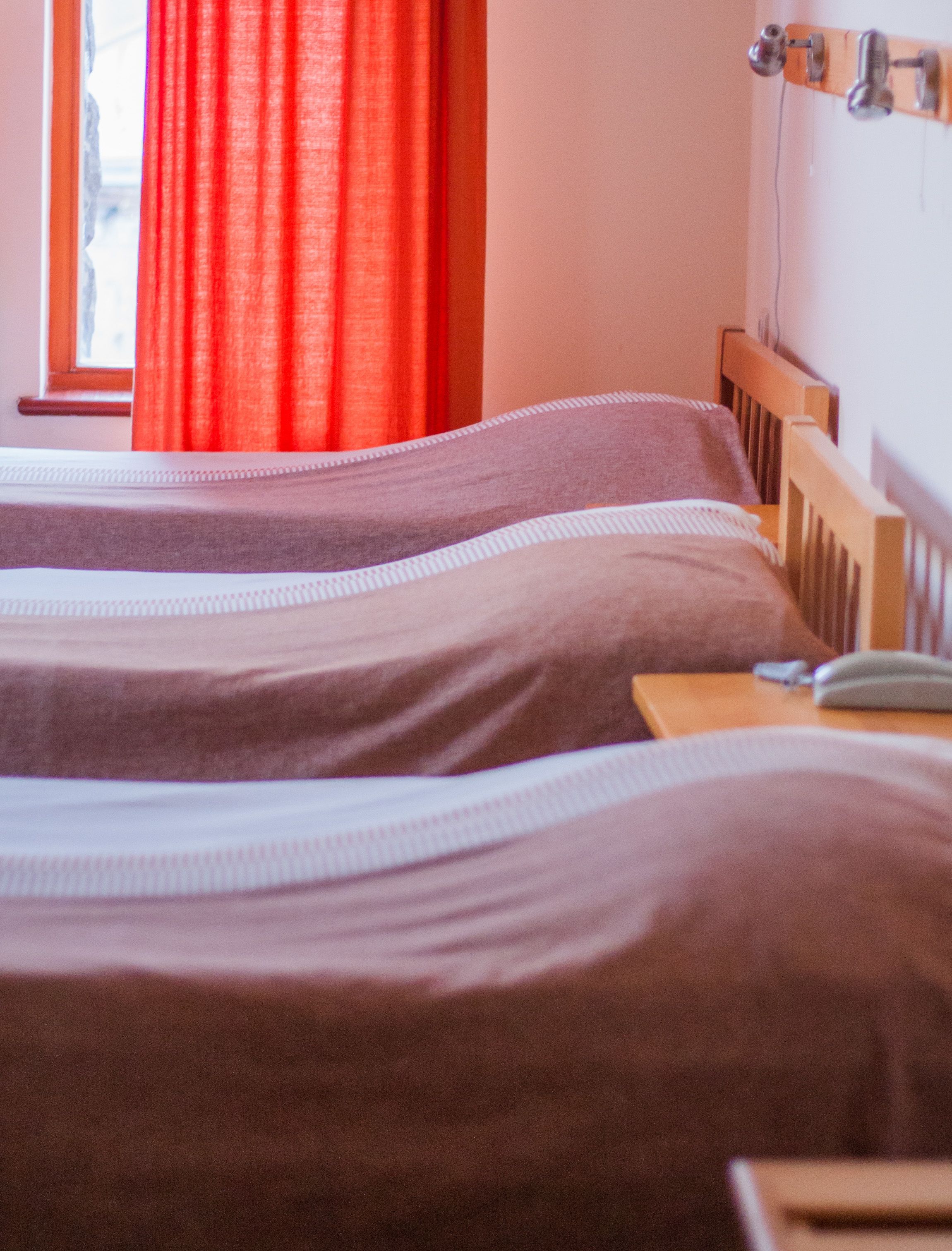 Una cama con cabecera de madera, una mesita de noche con lámpara y una ventana con cortinas rojas.