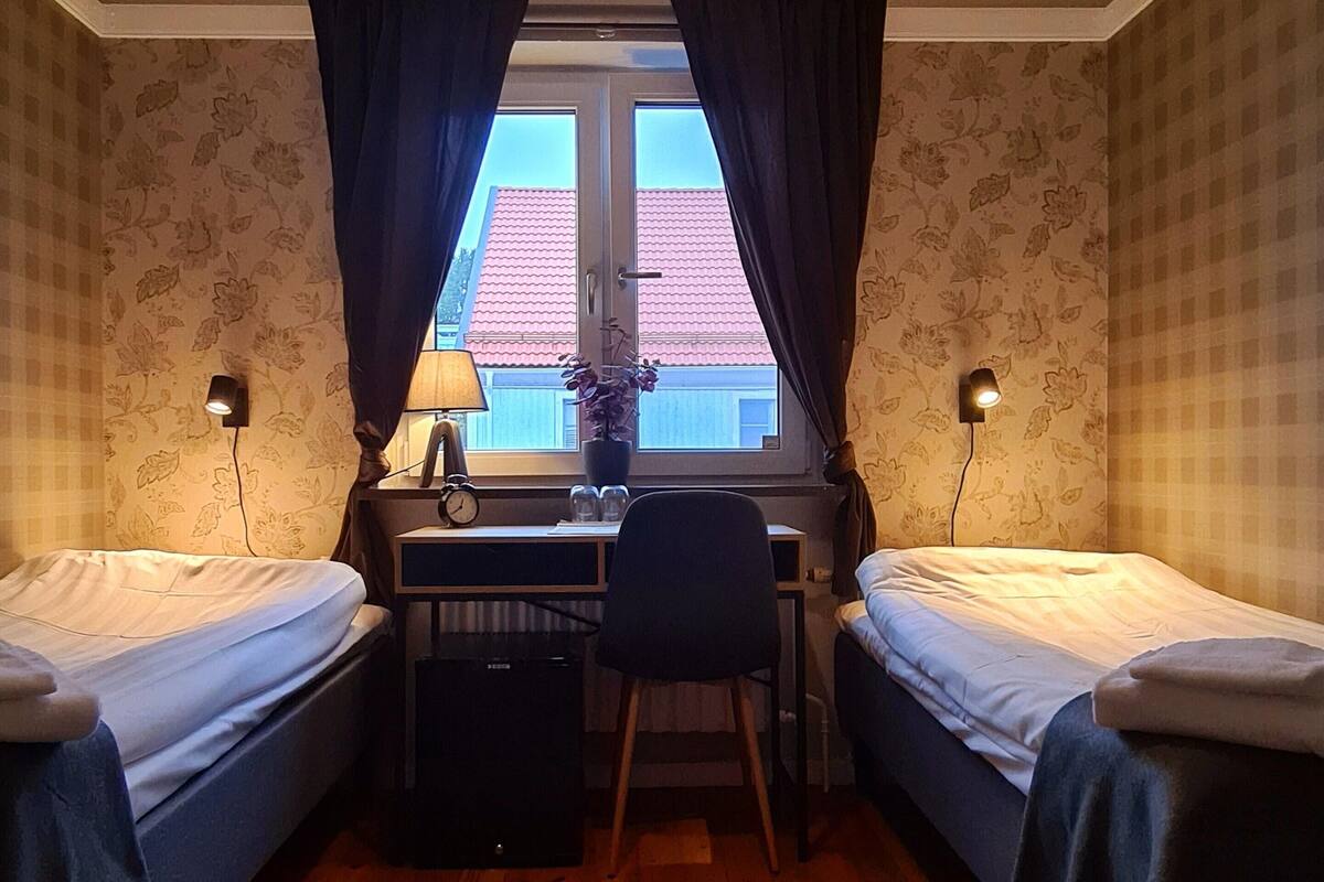 Une chambre avec deux lits simples, un bureau avec une chaise, une petite table avec une lampe et une fenêtre avec des rideaux.