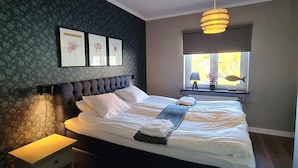 Een slaapkamer met een bed, nachtkastje, wanddecoratie en een raam met uitzicht.