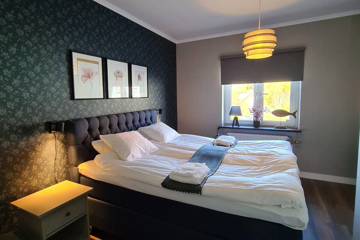 Une chambre à coucher comprenant un lit, une table de chevet, une œuvre d’art accrochée au mur et une fenêtre donnant sur un paysage.