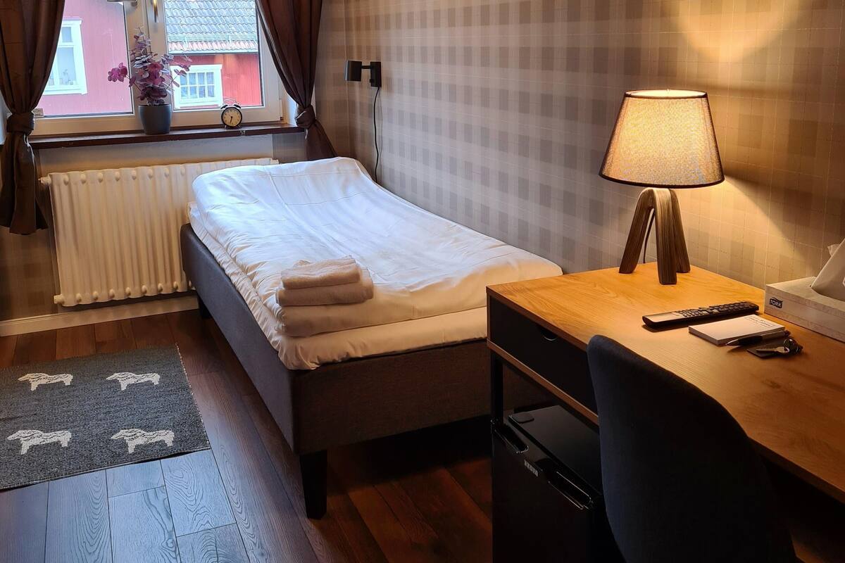 Une chambre d’hôtel avec un lit, un bureau, une chaise, une lampe et une fenêtre avec des rideaux.