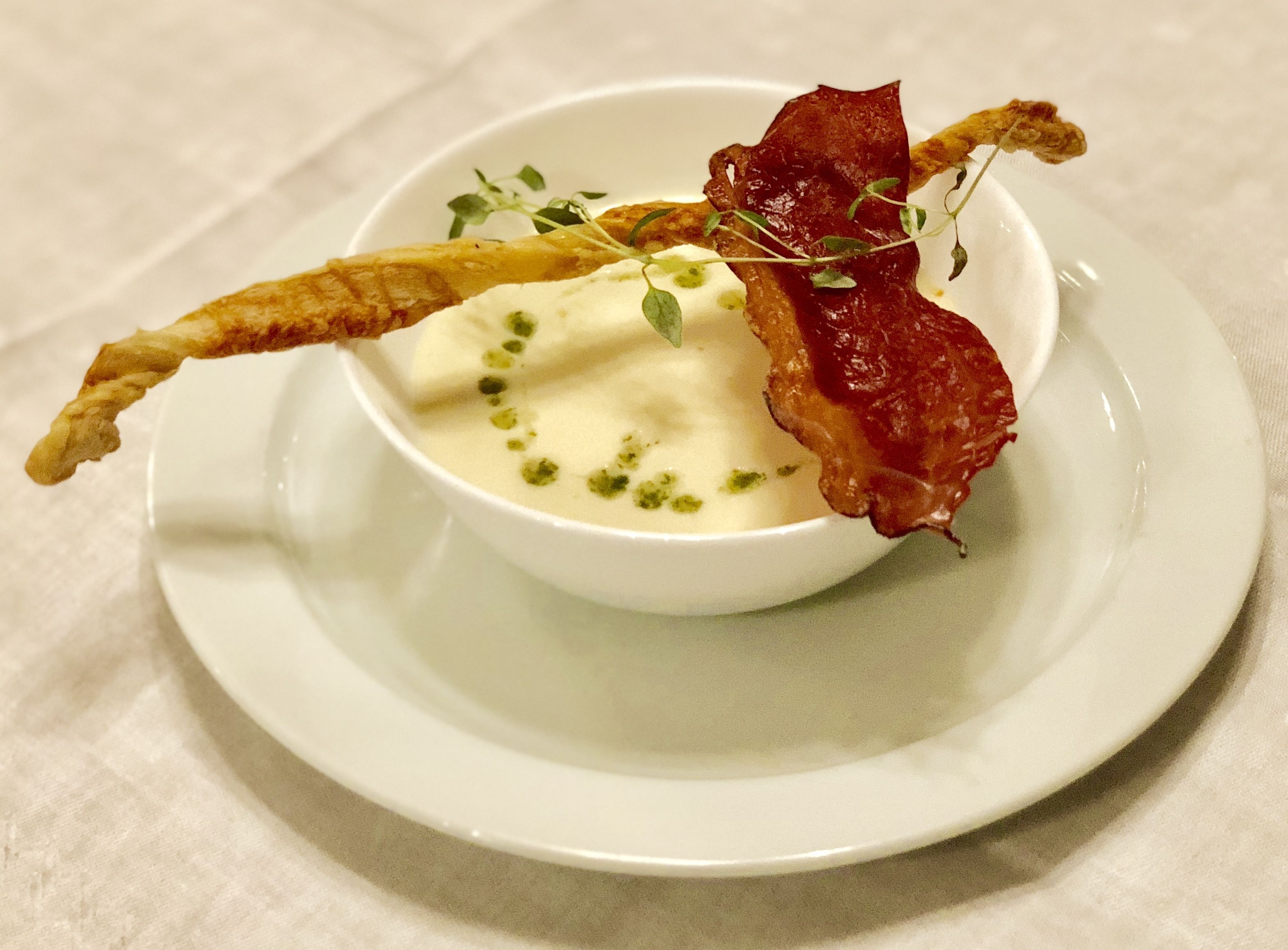 A white bowl of creamy soup with a garnish of green herbs, accompanied by crispy bacon strips and a fried item, served on a white plate.