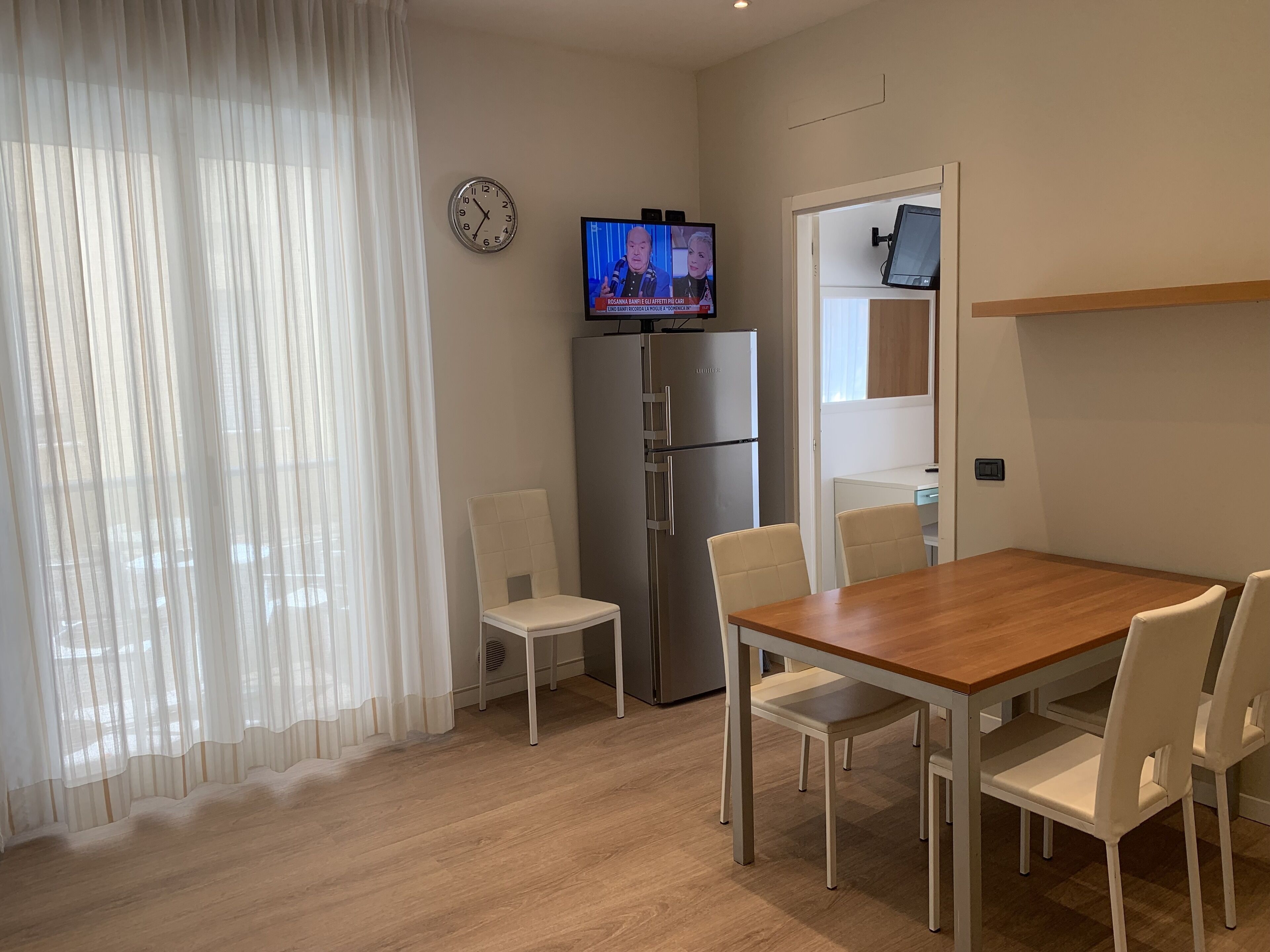 A modern kitchen with a refrigerator, a dining table with chairs, a clock, and a television displaying a news program.
