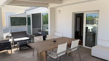 Un patio techado al aire libre con una mesa de madera y sillas blancas, rodeado de mobiliario de mimbre y un sofá blanco.