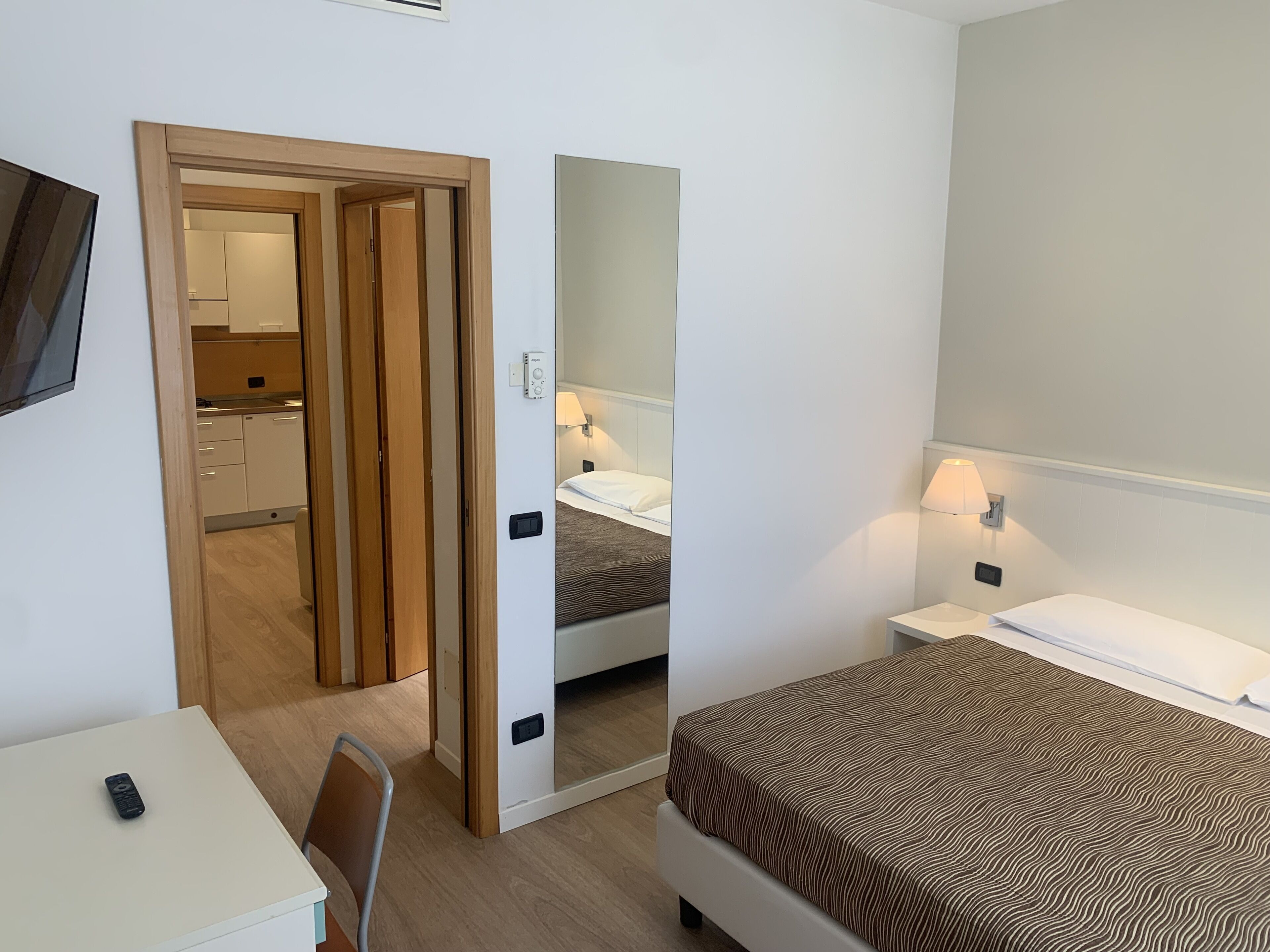 A hotel room with a bed, bedside table, and a small desk with a chair. There is a mirror on the wall and a door leading to a bathroom.