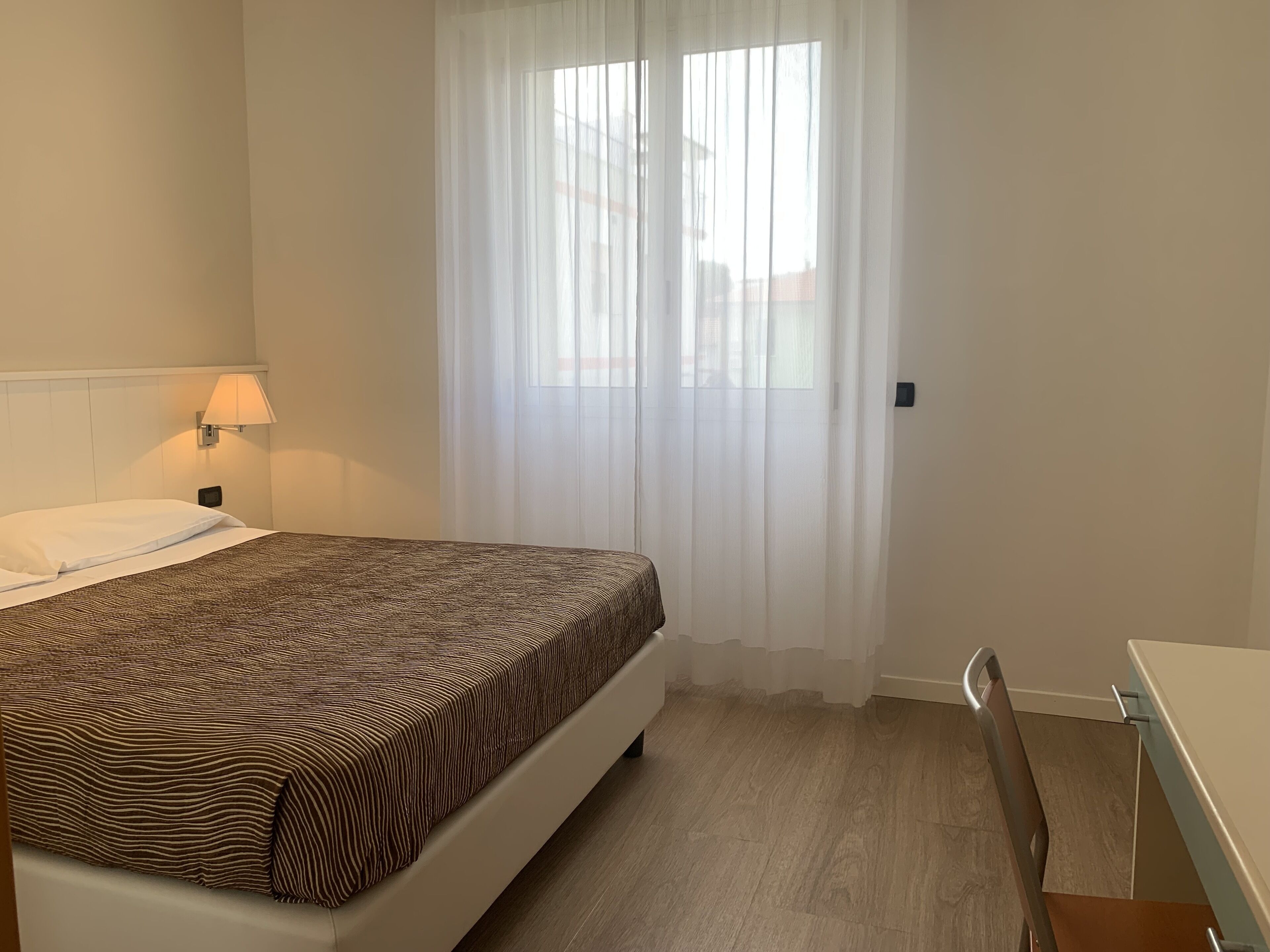 Een hotelkamer met een bed, een bedlampje, een raam met dunne gordijnen en een houten vloer.