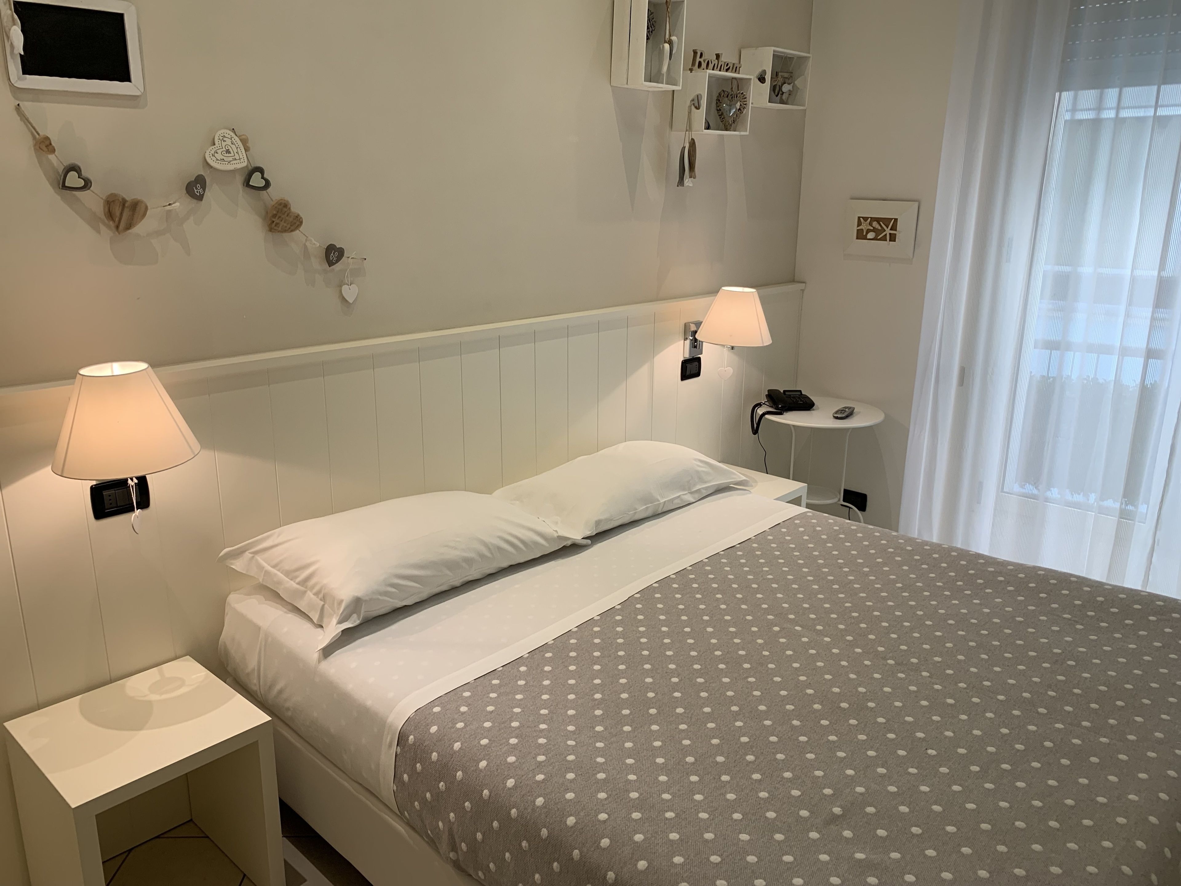 A neatly arranged hotel room with a bed, two bedside lamps, a small bedside table, and a wall-mounted shelf with decorative items.