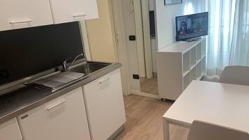 Una cocina moderna con armarios blancos, un lavamanos de acero inoxidable y un horno empotrado. Un comedor contiguo presenta una mesa y sillas blancas. Un televisor está montado en la pared y una puerta conduce a otra habitación.
